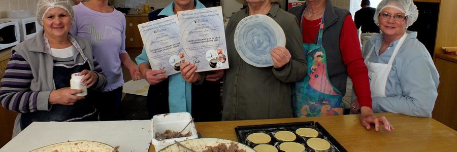 Onderskeidelik 3de en 4de van links is Suster Linda Meyer en Francia van Dyk met die sertifikaat en bord-embleem wat deur BADISA aan Gansbaai se Goue Hande toegeken is, saam met enkele lede by hulle wat besig is om pasteie te bak